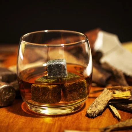 A glass of whiskey on the rocks in simple glass ware with whiskey stones in a carpentry work space with a hammer and saw with wood chips on top a wood desk.